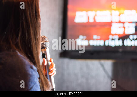 A woman singing at the karaoke bar holding a microphone in front of TV screen with lyrics. Stock Photo