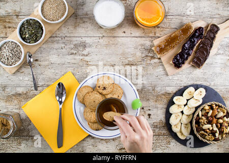A woman's hand soaking the biscuit in tea at a wooden table set for a sweet vegan breakfast shot from above with sliced banana, mixed dried fruit, veg Stock Photo