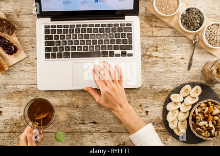 The habits of modern times: a woman shot from above on a wooden table, makes a healthy and vegan breakfast, but in front of the open laptop Stock Photo