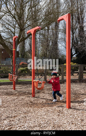 Two year old boy playing with swings in the park wearing a bobble hat and red coat Stock Photo