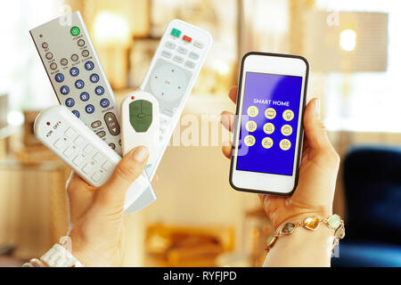 Closeup on remote controls in one hand and smartphone with smart home application in another hand of modern housewife at home. Stock Photo
