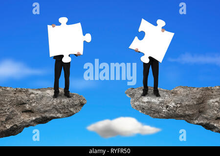 Businessmen holding two white jigsaw puzzles to connect on the cliff with sky background Stock Photo