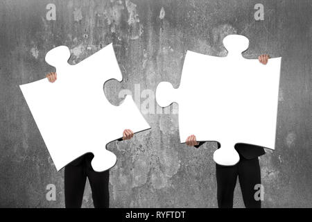 Two business people assembling blank white jigsaw puzzles with concrete wall background Stock Photo