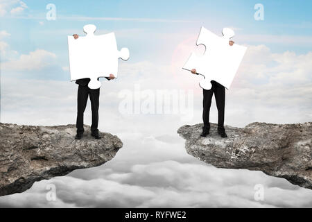 Businessmen holding two white jigsaw puzzles to connect on the cliff with cloudscape sunlight background Stock Photo