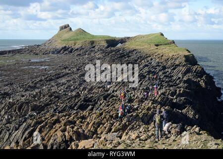 Hikers crossing the 'Low neck' from the Inner Head on the Worm's head to reach the 'Devil's Bridge' rock archway and the Outer Head, Rhossil, Wales. Stock Photo