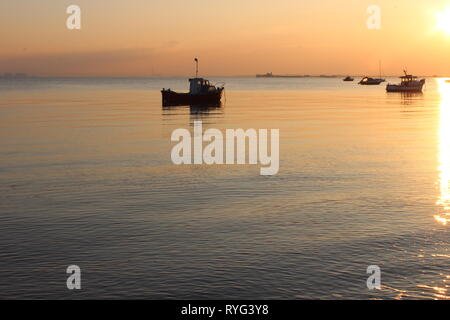 colour image of a sunset showing a trawler and other boats moored at low tide on calm water with the sun reflecting Stock Photo