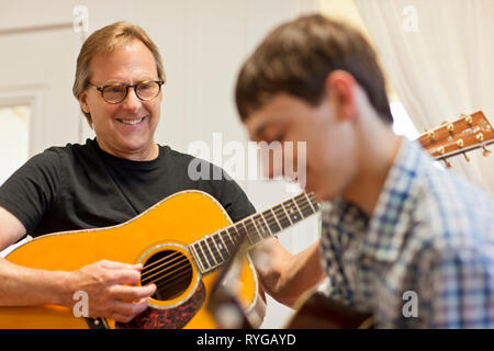 Mature man holding an acoustic guitar while watching his son play a guitar along with him. Stock Photo