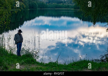 Man in hat is silhouetted as he stands by the side of the water near sunset in the lake district of northeastern Poland near Suwalki Stock Photo