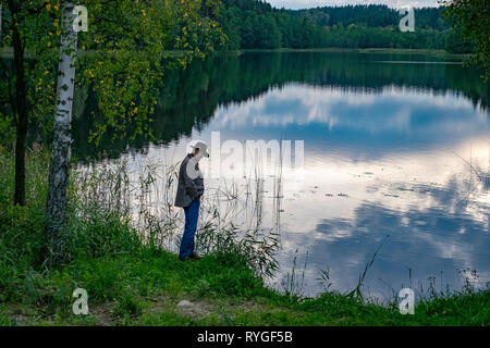 Man in hat is silhouetted as he stands by the side of the water near sunset in the lake district of northeastern Poland near Suwalki Stock Photo