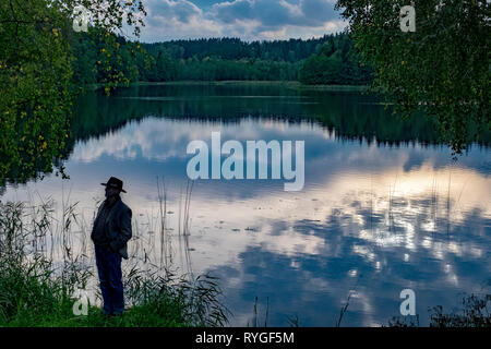 Man in hat is silhouetted as he stands by the side of the water near sunset in the lake district of northeastern Poland near Suwalki Stock Photo