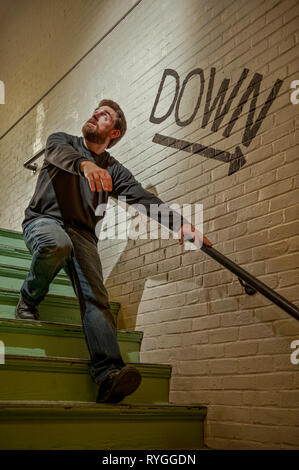 Bearded man scrambles down a staircase in fear or terror with a Down arrow guiding his path Stock Photo