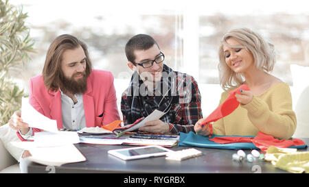 Smiling tailor businesswoman sitting at sewing machine and making handmade clothing while marketing assistant working on new projec Stock Photo