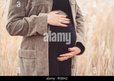 Pregnant unrecognizable woman in black dress standing in reeds and holding her belly. Stock Photo