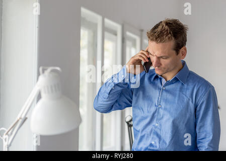 Serious young businessman standing by the office windows engaged in an important conversation using his smart phone. Stock Photo