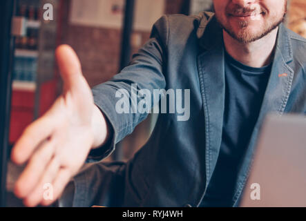 Smiling attractive adult successful bald bearded man in suit with laptop giving handshake, hand of help, greeting at cafe Stock Photo