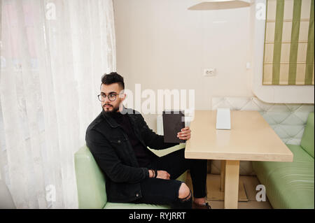 Arab man wear on black jeans jacket and eyeglasses sitting in cafe, read book. Stylish and fashionable arabian model guy. Stock Photo