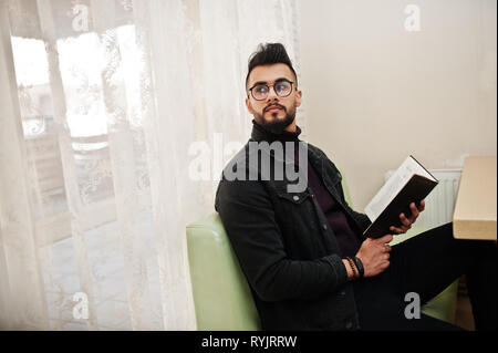 Arab man wear on black jeans jacket and eyeglasses sitting in cafe, read book. Stylish and fashionable arabian model guy. Stock Photo