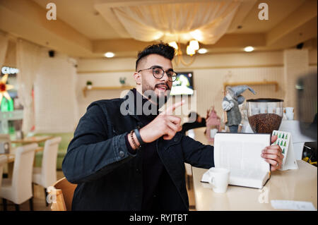 Arab man wear on black jeans jacket and eyeglasses in cafe drink coffee at bar with book. Stylish and fashionable arabian model guy. Talking to barman Stock Photo