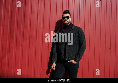 Fashion Arab man wear on black jeans jacket and sunglasses posed against red steel wall background. Stylish and fashionable arabian model guy. Stock Photo