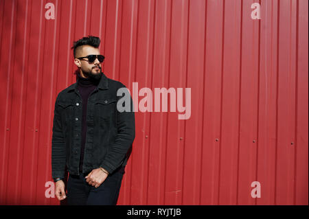 Fashion Arab man wear on black jeans jacket and sunglasses posed against red steel wall background. Stylish and fashionable arabian model guy. Stock Photo