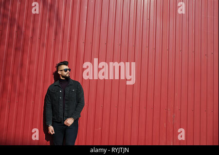 Fashion Arab man wear on black jeans jacket and sunglasses posed against red steel wall background. Stylish and fashionable arabian model guy. Stock Photo