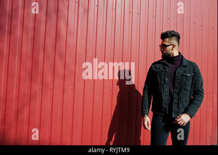 Fashion Arab man wear on black jeans jacket and sunglasses posed against red steel wall background. Stylish and fashionable arabian model guy. Stock Photo