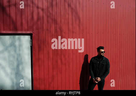 Fashion Arab man wear on black jeans jacket and sunglasses posed against red steel wall background. Stylish and fashionable arabian model guy. Stock Photo