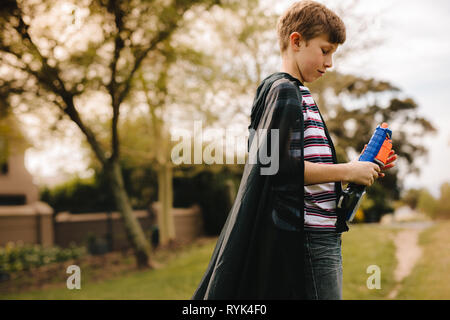 Side view of a young boys wearing a cape playing with toy gun outdoors. Boy with cape pretending to a superhero. Stock Photo