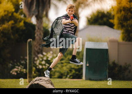 Young boy wearing cape jumping over a log with a toy gun. Boy pretending to be superhero fighting with toy gun in backyard. Stock Photo