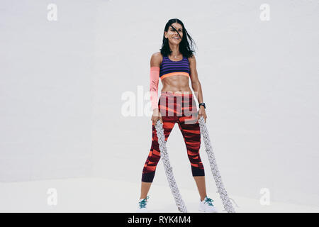 fitness woman standing holding two battle ropes during workout. Woman in fitness wear working out using battle ropes. Stock Photo