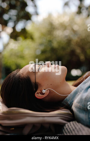 Close up of a young woman lying on ground in a park wearing sunglasses. Woman relaxing outdoors listening to music. Stock Photo
