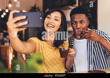 Woman taking selfie with friend making funny expressions while drinking coffee. Friends taking selfie with smart phone at cafe. Stock Photo
