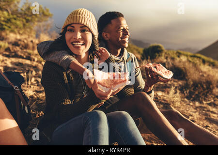 Two friends having pizza sitting on mountain trail. Man and woman on hiking trip eating pizza. Stock Photo