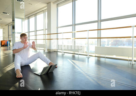Young male person learning new movements with tutorial using laptop. Male person sitting in white shirt and pants. Concept of Internet sources for sel Stock Photo