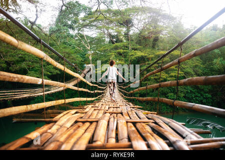 Back view of young woman on suspension wooden bamboo bridge across Loboc river in jungle. Vacation on tropical island. Bohol, Philippines Stock Photo