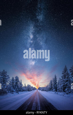 Road leading towards colorful sunrise between snow covered trees with epic milky way on the sky Stock Photo