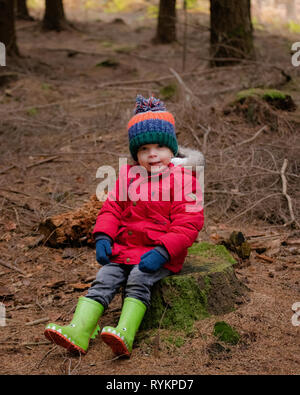 Two year old toddler sat on tree stump in the forest during autumn wearing a red coat, wellies and a bobble hat Stock Photo