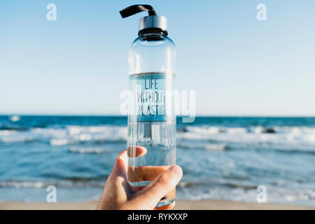 closeup of a caucasian man holding a glass reusable water bottle with the text life without plastic written in it, on the beach, with the ocean in the Stock Photo