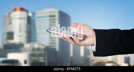 Businessmans Hand Holding Smartphone With Business City and Corporate Buildings In Background Stock Photo