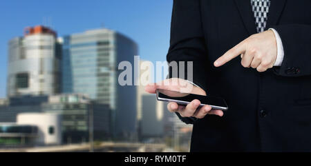 Businessman Holding Smartphone in Hand And Pointing Index Finger At The Phones Screen With Business City and Corporate Buildings In Background Stock Photo