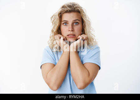 Close-up shot of insecure and silly scared cute female with blond hair and blue eyes clench teeth shaking from fear holding hands on jawline, worry Stock Photo