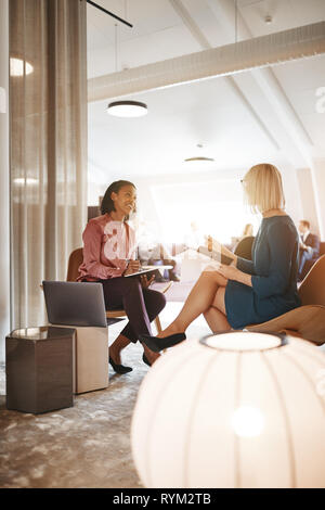 Two smiling young businesswomen sitting together in a modern office working on a laptop and going over paperwork Stock Photo