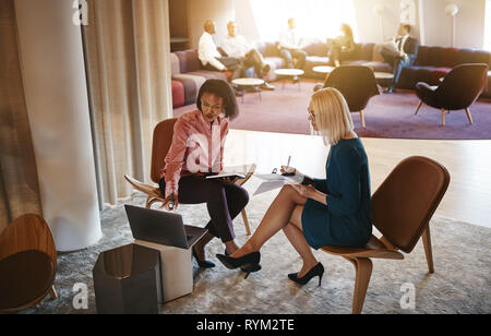 Two smiling businesswomen sitting together in a modern office working on a laptop and going over documents Stock Photo