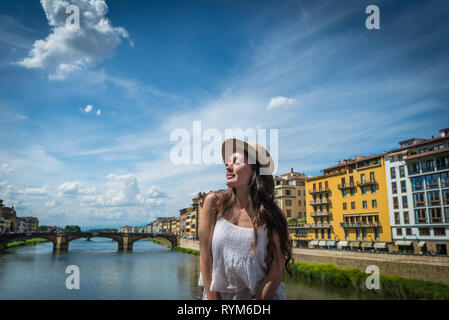 Young woman enjoys holidays in Florence, Italy. Beautiful girl with smile and closed eyes in old town sits with view on river, sky, houses and bridge. Stock Photo