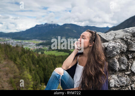 Female traveler sits on peak of mountain with closed eyes on background of valley, forest and village. Young Kazakh girl enjoying day in Alps, Austria Stock Photo