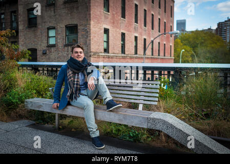 Young elegant man relaxing with view on buildings and trees in High Line park in New York City, USA. Handsome guy sits on bench and looking in camera. Stock Photo