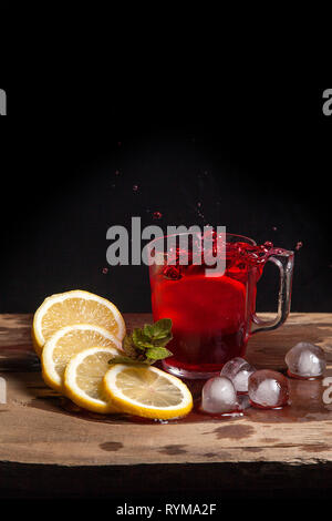 Summer iced drink - hibiscus cold tea with ice, lemon and mint.  Big splash with drops of hibiscus cold tea from the glass of beverage with fallen lem Stock Photo