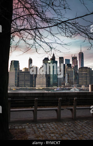Scenic view from Brooklyn on Manhattan skyscrapers in evening time in New York, USA. Benches on riverside on background of city downtown and sunset. Stock Photo