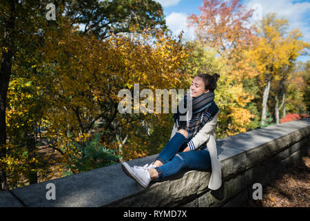 Young smiling woman resting on background of yellow trees and leaves. Happy elegant girl sits alone in park. Sunny autumn day in New York city, USA. Stock Photo