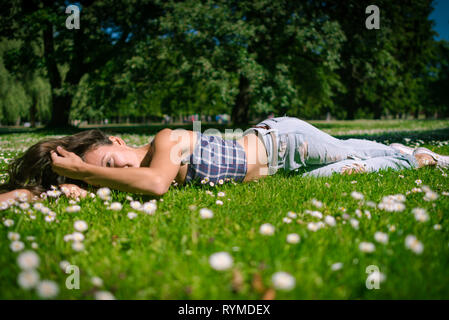 Young joyful woman lies on green grass. Happy girl is relaxing at chamomile field in warm summer day on background of big trees in park. Stock Photo